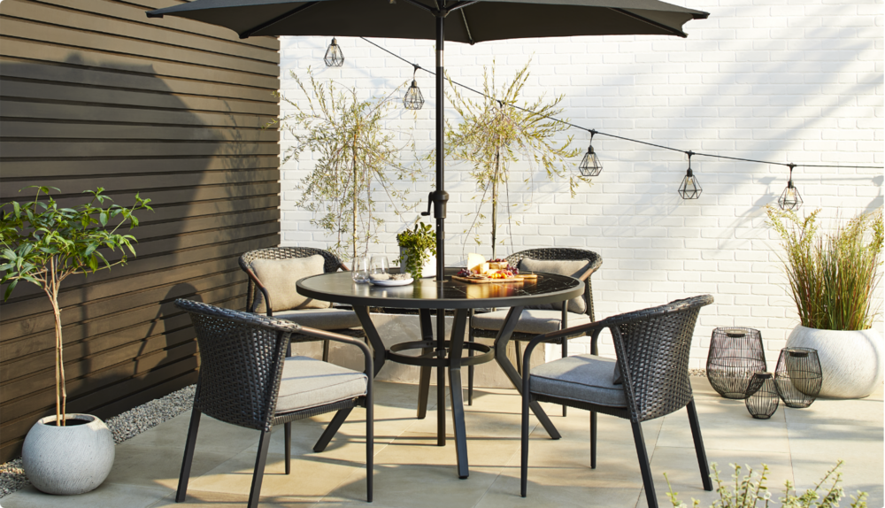 CANVAS Jasper Dining Collection displayed on a backyard deck featuring a large umbrella, four wicker dining chairs, a round table, outdoor string lights, decorative lanterns and plants. 