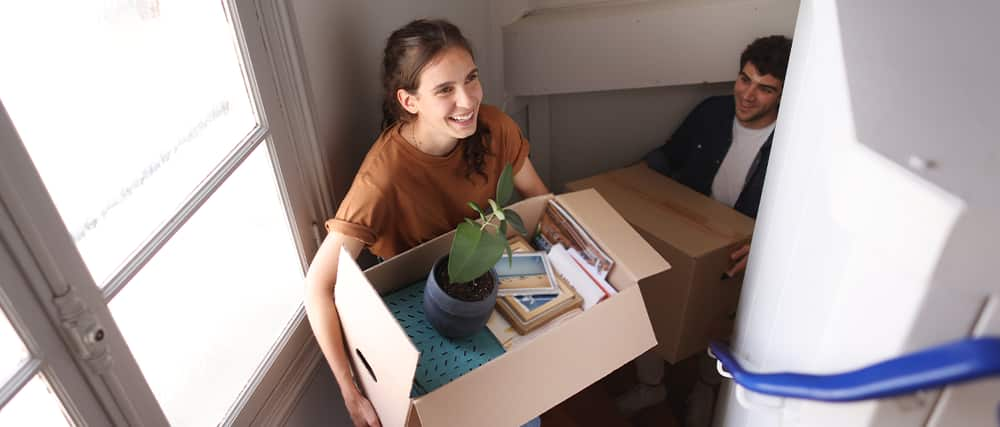 Person packing away Christmas decorations in a cardboard box with bubble wrap. Also pictured, a roll of packing tape, tape gun, roll of bubble wrap, and packing tape with dispenser.