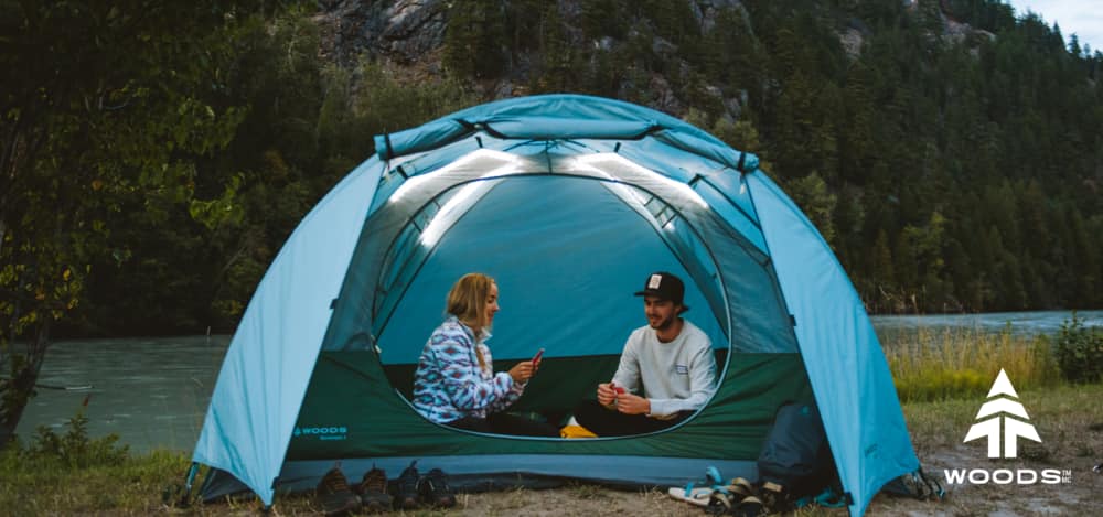 Une tente Woods ILLUMINATE ouverte avec ses bandes lumineuses allumées, et deux jeunes adultes jouant aux cartes à l'intérieur.