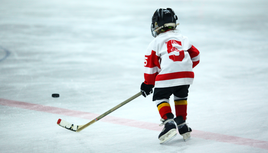 Un jeune joueur de hockey en chandail d’entraînement blanc frappe vers le gardien de but à l’intérieur d’une patinoire de hockey.