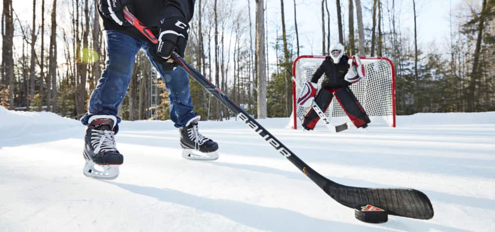 Two youths are playing recreational hockey in a woodsy outdoor rink using a Canadian Tire hockey puck and Bauer hockey stick.