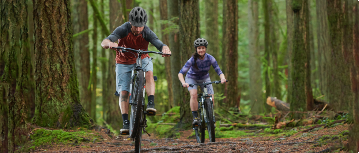 Two people ride Raleigh mountain bicycles along a wooded trail.