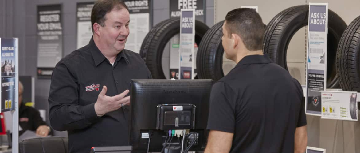 A Canadian Tire auto service employee speaks with a customer next to a rack of tires.