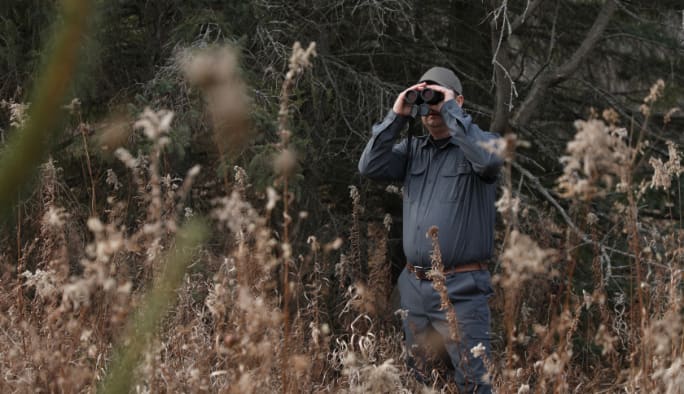 A hunter in dark clothes looks through binoculars in a wooded setting.