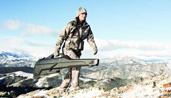 A hunter in camoflauge carries a rifle case up the side of a mountain.