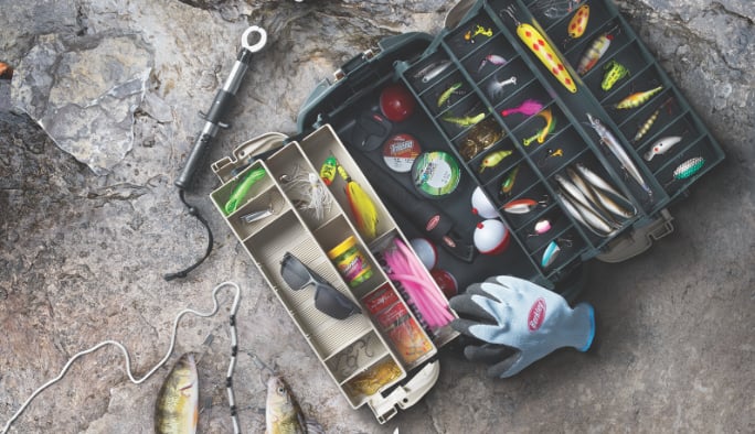 Une coffre à pêche ouverte remplie de leurres de pêche placée sur des rochers à côté d’un gant de pêche à la mouche et de deux poissons pêchés.