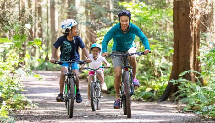 Un adulte et deux enfants portant des casques, faisant du vélo sur un sentier boisé.