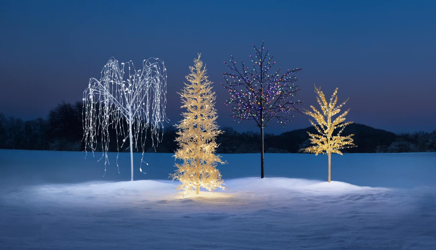 A range of CANVAS Outdoor Pre-Lit Trees on a snowy lawn including the Twinkling Willow Tree and Twig Tree.