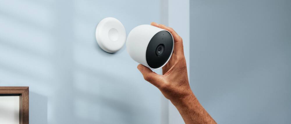 A hand reaches to install a smart camera high on a pale blue wall.