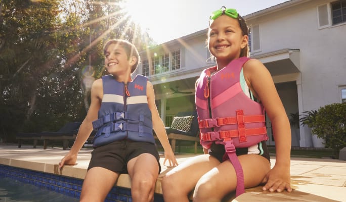 Two kids sitting on edge of pool with life jackets 