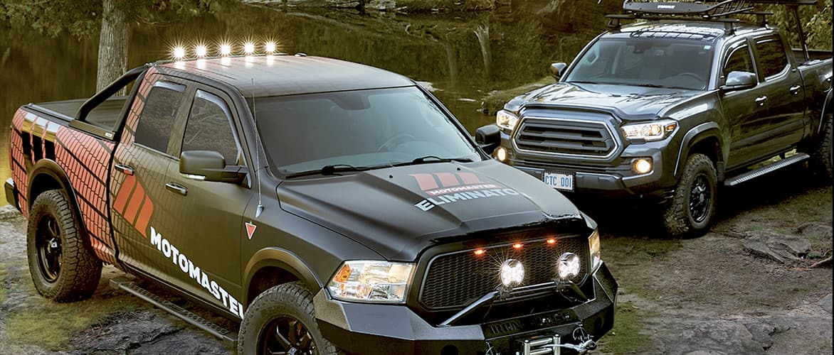 Two black pickup trucks sit parked near a lake in the woods.