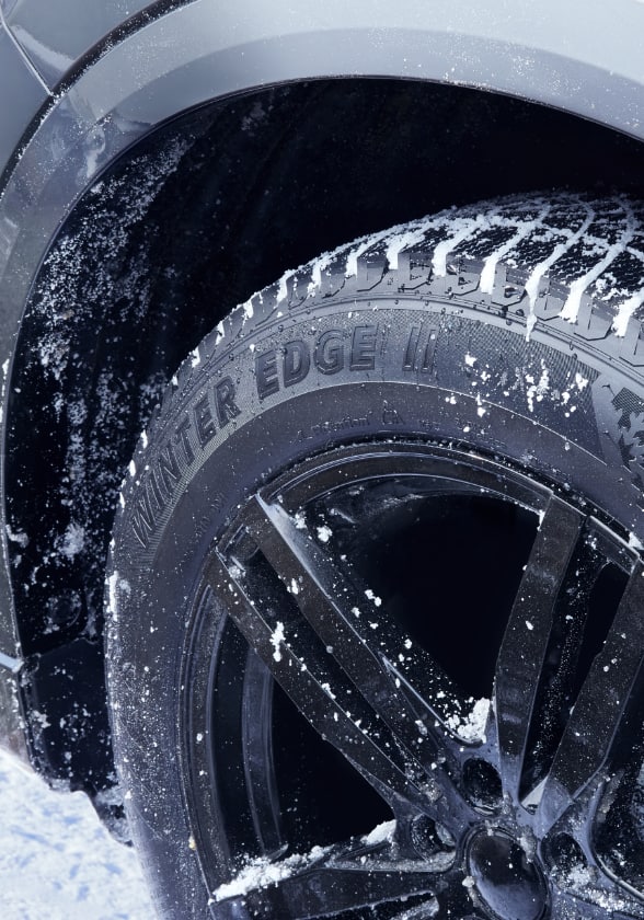 A MotoMaster Winter Edge II tire mounted in the wheel well of a car on a snowy road.