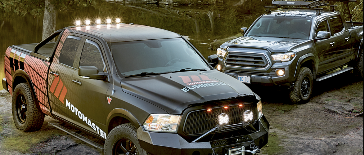Two black pickup trucks fitted with aftermarket accessories sit parked in a wooded setting.