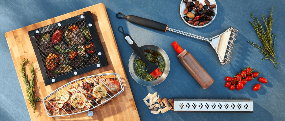 Une brosse en serpentin Vida par PADERNO et d’autres accessoires pour barbecue sur une table avec des aliments grillés.