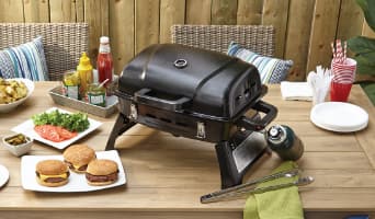 A MASTER Chef Portable Gas Grill on a patio table with cooked burgers and other foods. 