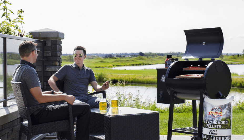 Two adults sitting next to Pit Boss 700FB1 Series Pellet Grill on a lakeside patio.