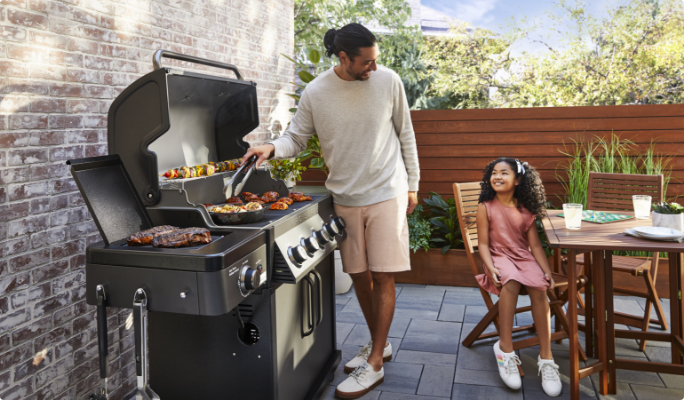 A man and a child chatting on a backyard patio while the man grills food on a gas BBQ.