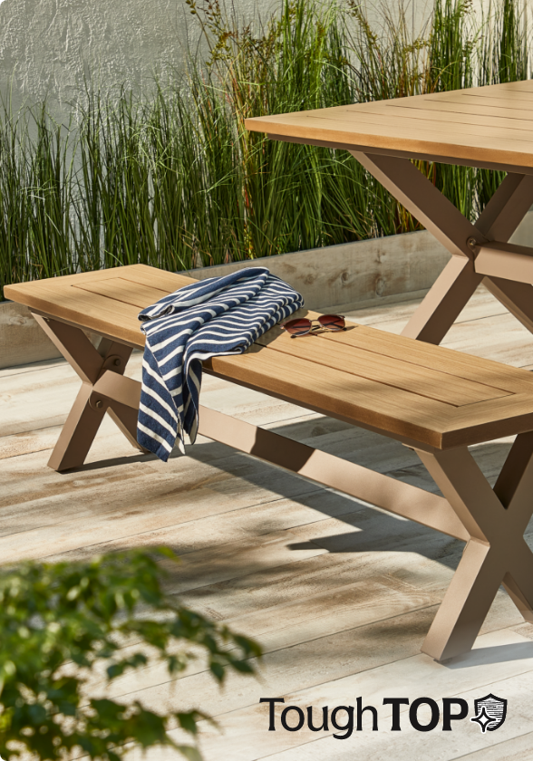 Un banc à dîner CANVAS Belwood sur une terrasse d’arrière-cour en bois avec des articles décoratifs en bois, des lunettes de soleil et une écharpe rayée sur le banc.