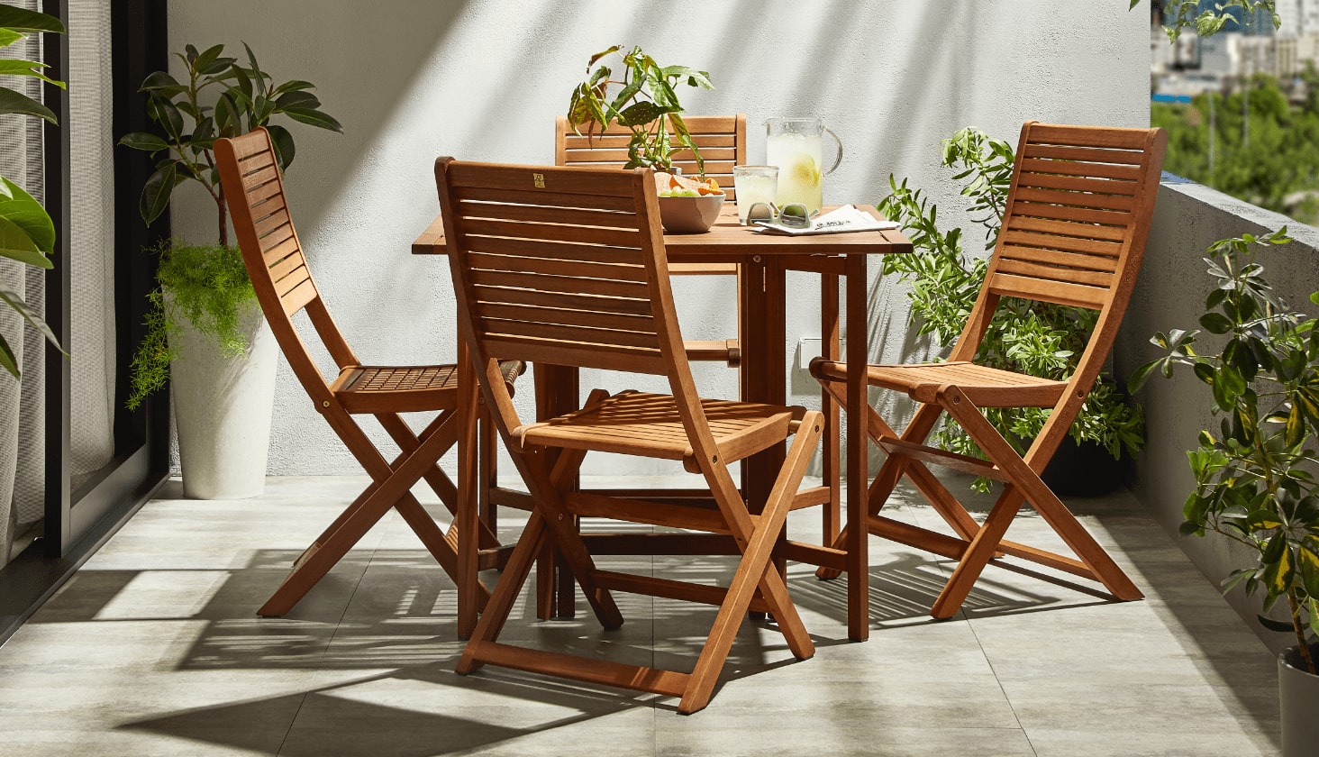 A wooden CANVAS Sherbrooke Folding Dining Set on a balcony with plants and snacks.