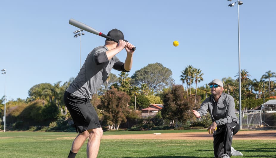An adult with a softball bat gearing up to hit a ball in mid-air. 