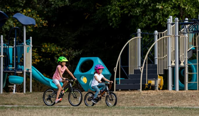 Two children in bike helmets, cycling past a playground. 