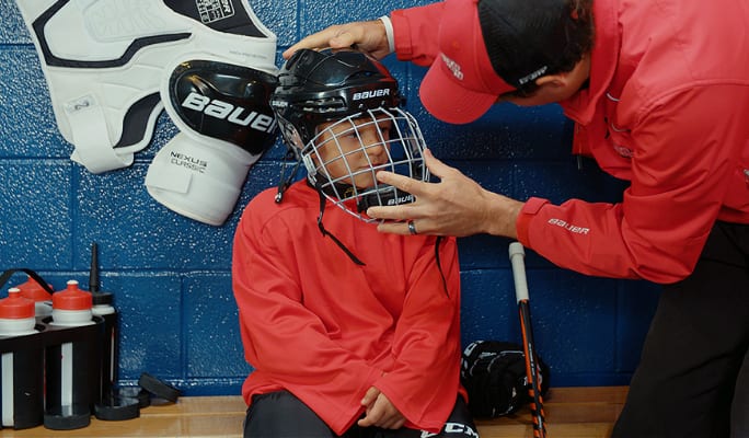 An adult helping a child put on a hockey helmet in a locker room. 