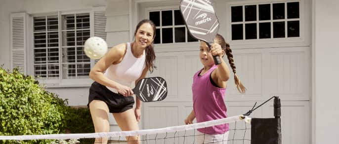 A woman and girl play pickleball in a driveway.