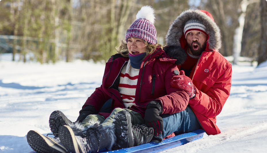 Two adults in winter parkas riding a sled in the snow. 