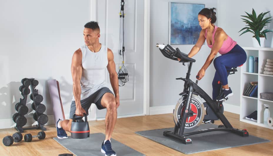 Two people exercise at home with weights and a stationary bike.