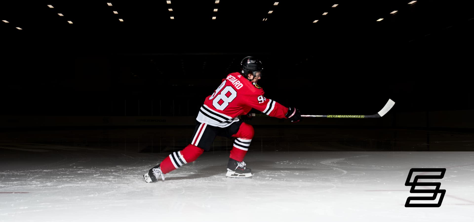  Connor Bedard in a Blackhawks uniform on a rink taking a shot with a hockey stick. 