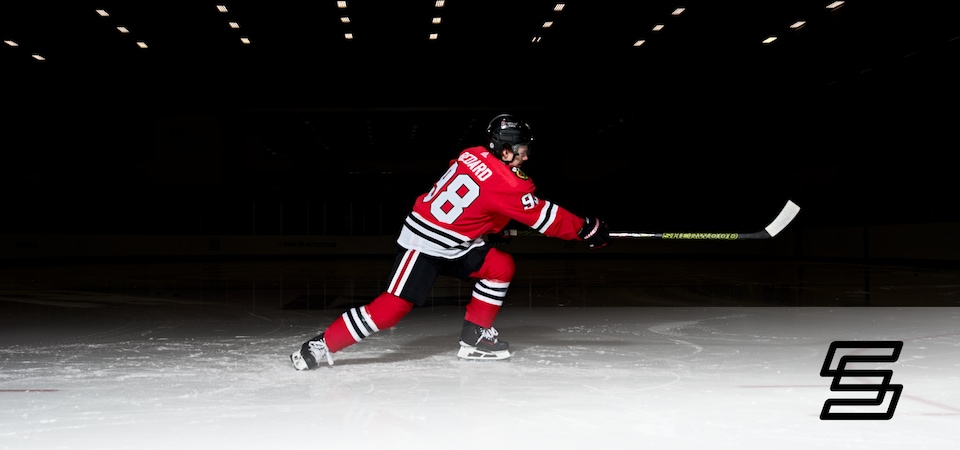  Connor Bedard in a Blackhawks uniform on a rink taking a shot with a hockey stick. 