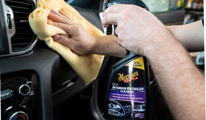 A person holding a bottle of car interior cleaner while wiping a vehicle’s dash board with a cloth.