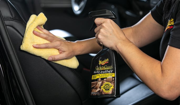 A person holding a bottle of leather cleaner while wiping a black leather car seat with a cloth.