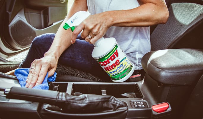 A person holding a bottle of multi-purpose cleaner while wiping a vehicle’s centre console with a cloth.