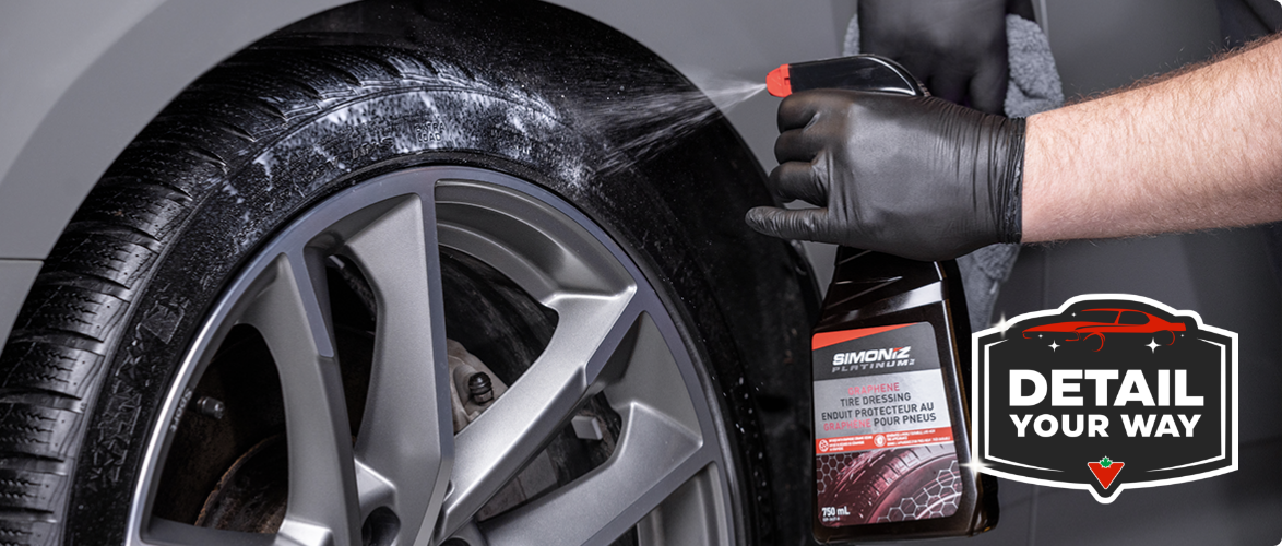 A hand wearing a black nitrile glove sprays SIMONIZ Platinum Graphene Tire Shine on a car tire.