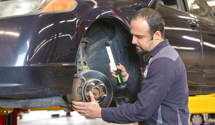 A technician holds a work light while inspecting the brakes of a car positioned on a hoist lift.