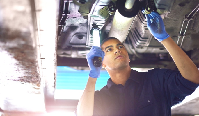 A technician holds a work light while inspecting the undercarriage of a car.
