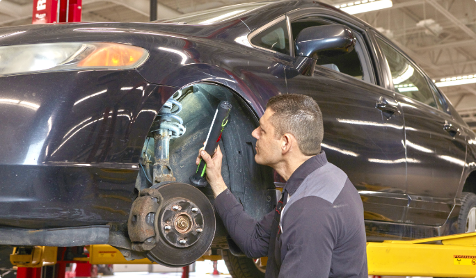 A technician holds a work light while inspecting the struts of a car positioned on a hoist lift.