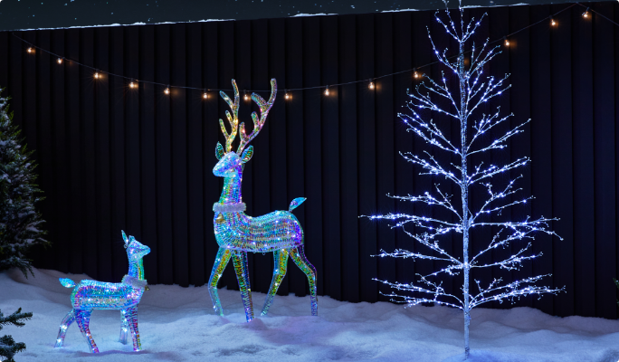 Des décorations de rennes scintillants CANVAS avec un arbre gemmé illuminé dans une cour enneigée. 