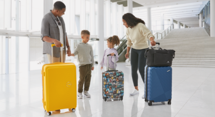Deux adultes et deux jeunes enfants à l'aéroport portant des bagages et des sacs de voyage American Tourister et Outbound.