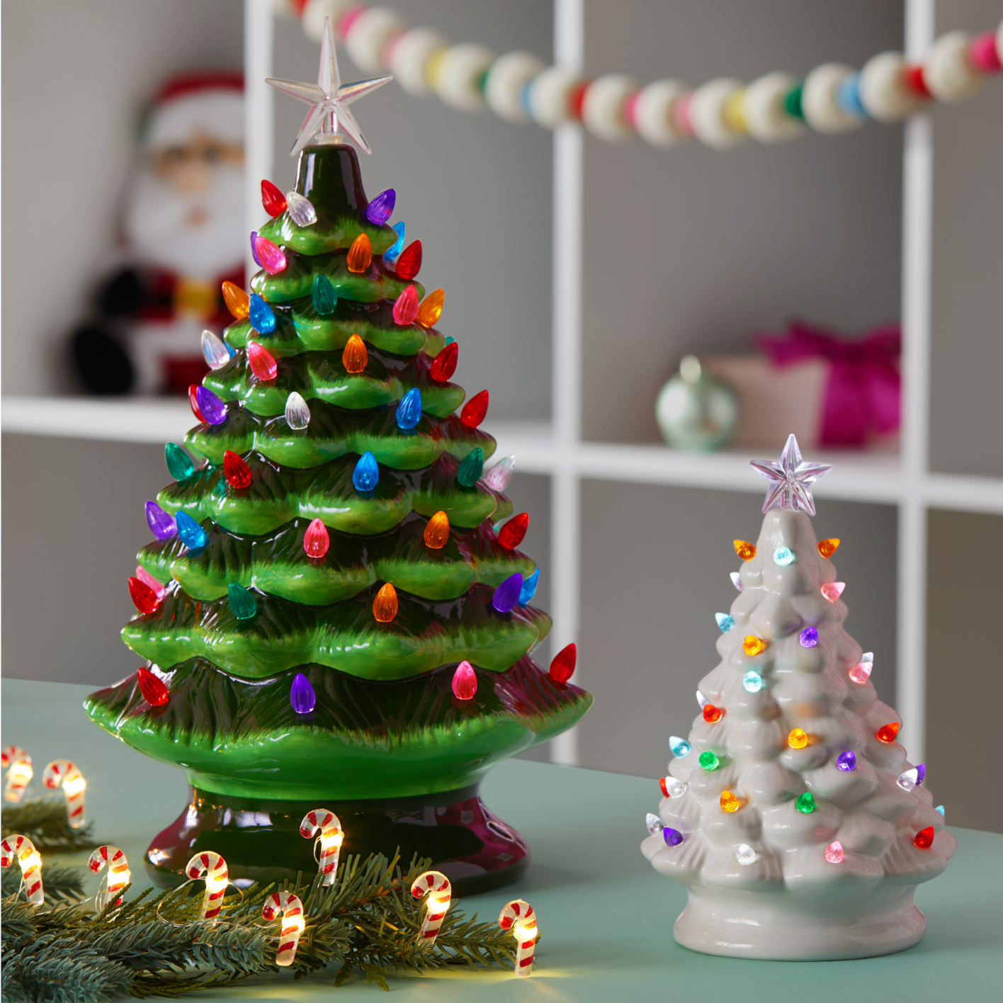 A green For Living ceramic tree, a white For Living ceramic tree and CANVAS candy cane string lights on a table. 