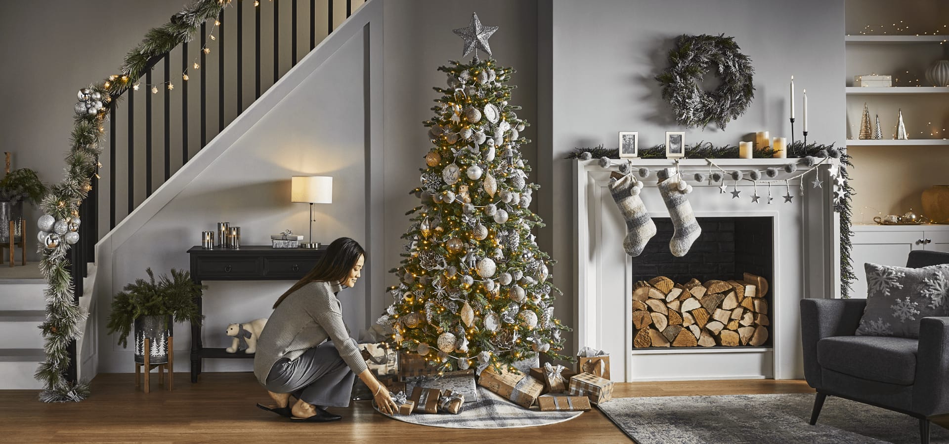 A woman putting wrapped gifts under a tree in a living room filled with CANVAS Silver Frost Christmas decor. 