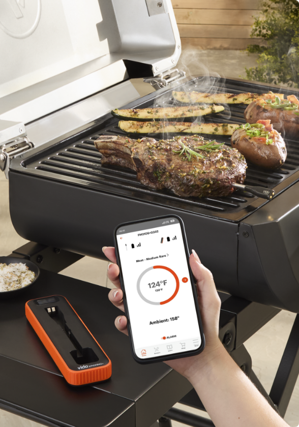 A VIDA by PADERNO Smart Thermometer on the side table of an open grill that is cooking steak and vegetables.