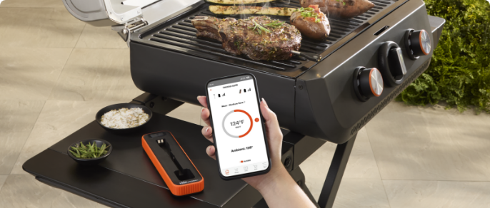 A VIDA by PADERNO Smart Thermometer on the side table of an open grill that is cooking steak and vegetables.