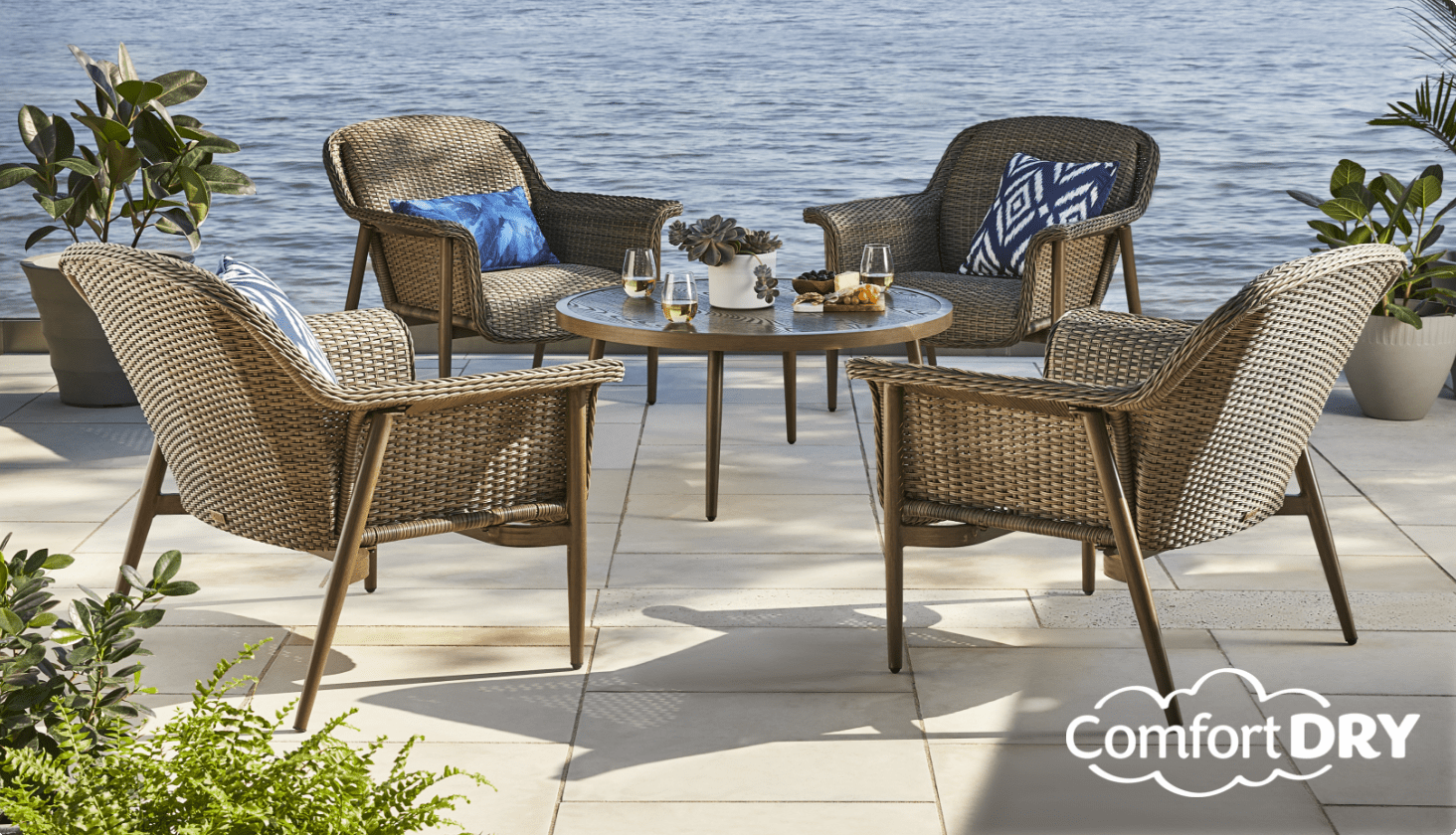 Quatre fauteuils club CANVAS Hunter et une table basse ronde avec des boissons sur une terrasse au bord de l’eau. 