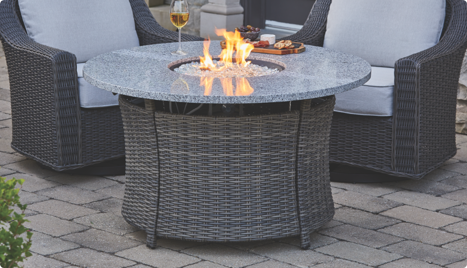 Deux fauteuils en osier à côté d’une table avec foyer au gaz CANVAS Summerhill allumé sur une terrasse.  