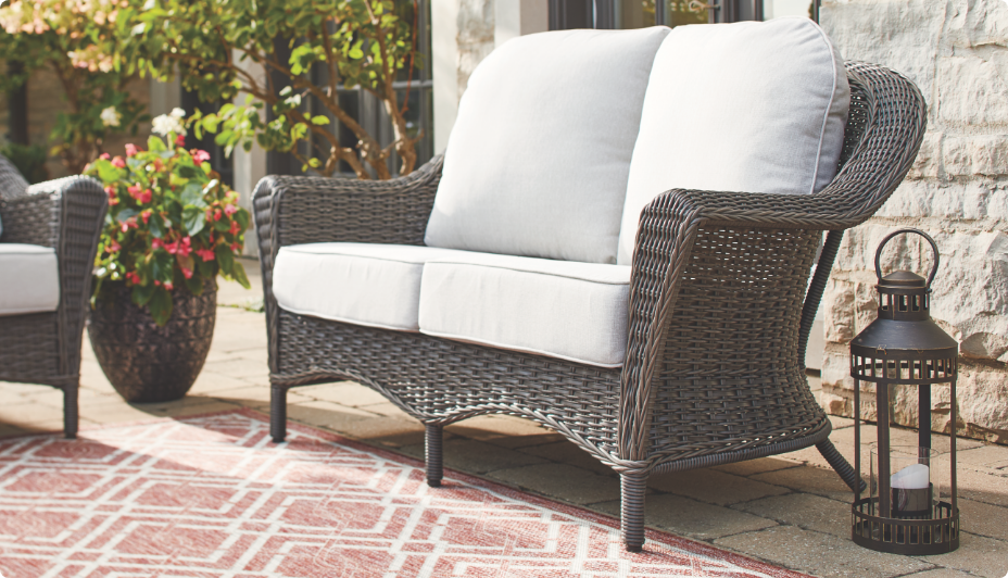 A wicker CANVAS Summerhill Loveseat with white cushions on a patio.   