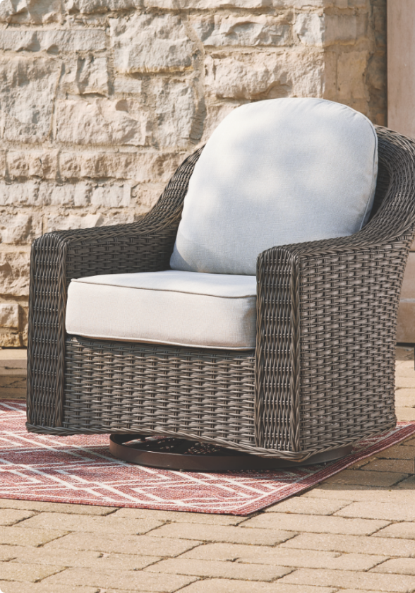 A wicker CANVAS Summerhill Swivel Glider Armchair with white cushions on a patio.