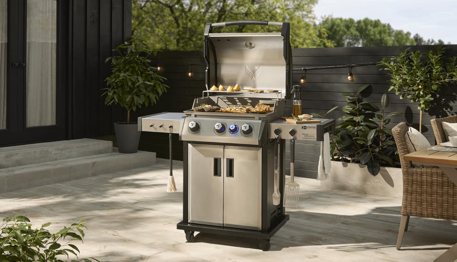 A Vermont Castings 3-Burner BBQ grilling various foods next to a patio table on a back patio.