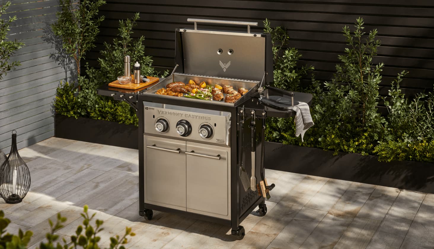 A Vermont Castings 2-Burner Griddle cooking food on a deck surrounded by various greenery.